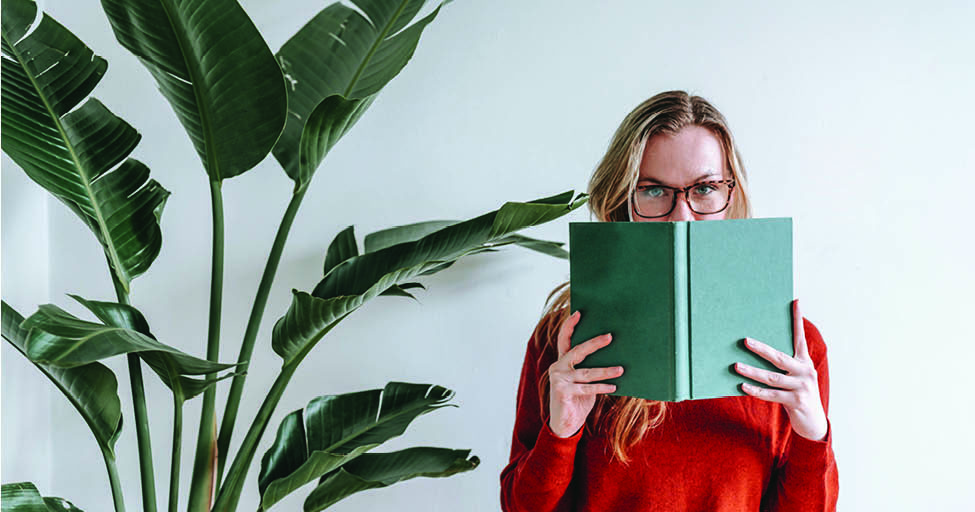 person holding book to their face by big plant
