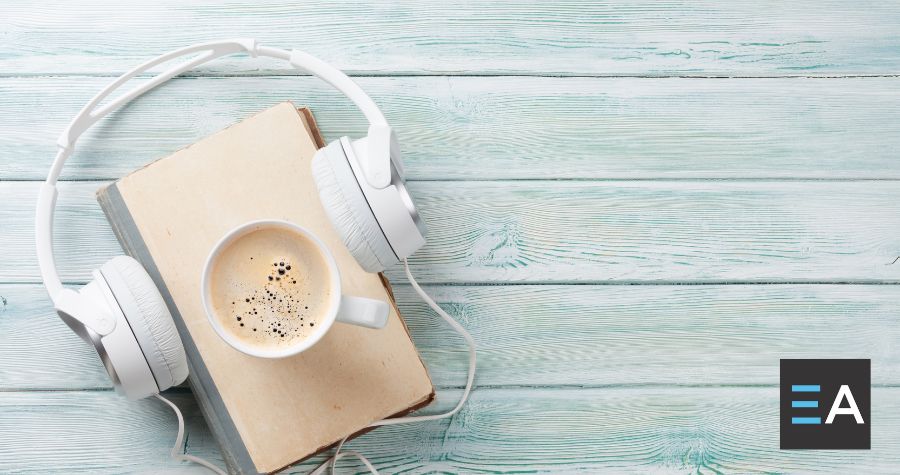 A full coffee mug on top of a book with headphones around it on a light colored wooden table