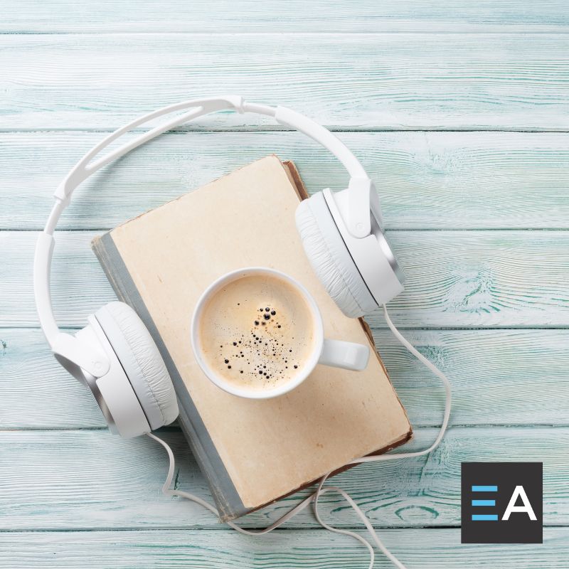 A full coffee mug on top of a book with headphones around it on a light colored wooden table