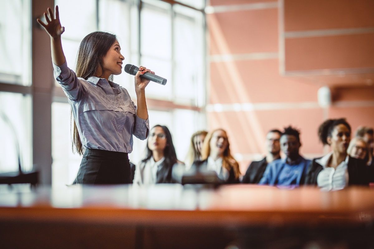 A presenter speaks into a microphone in front of an interested audience