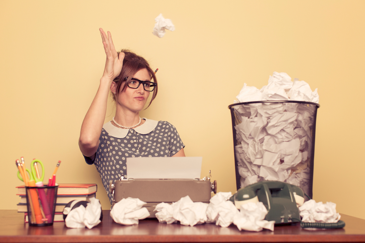A person using a typewriter at a desk tosses a crumpled piece of paper into a wastebasket