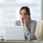 A person looking at a laptop with their hands on their face in a shocked expression