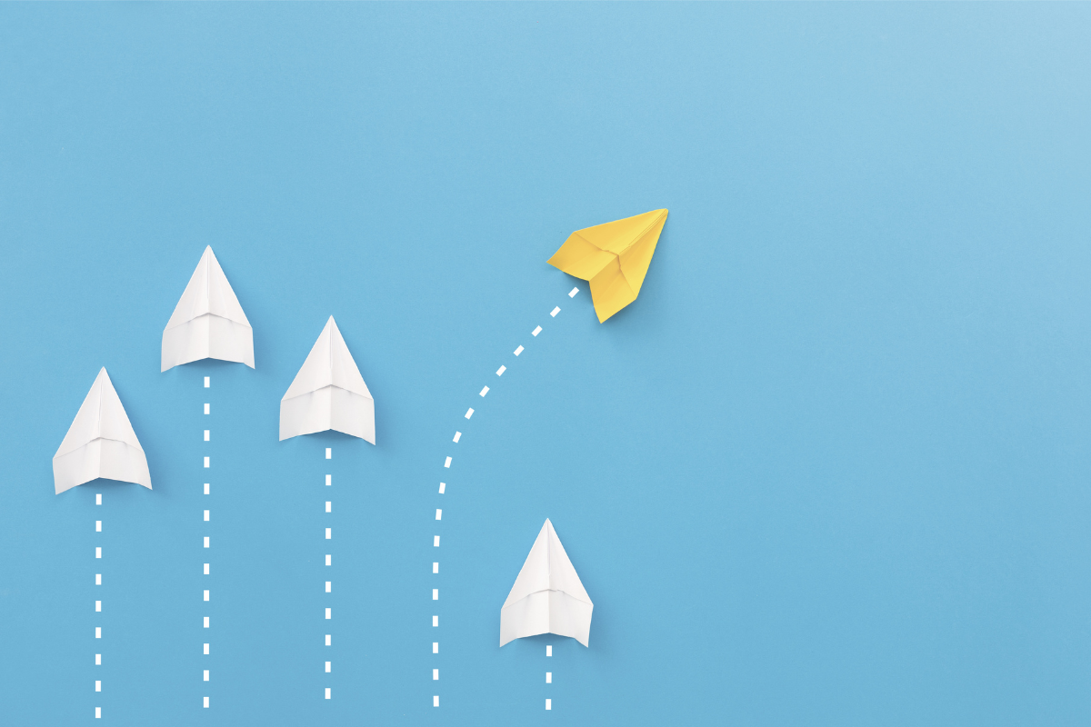 A yellow paper airplane diverges from the path that four white paper airplanes are following