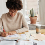 Busy accountant with curly dark hair, dressed casually, works on financial report at home, writes graphics and schemes, sits at desktop with coffee, documents and cell phone