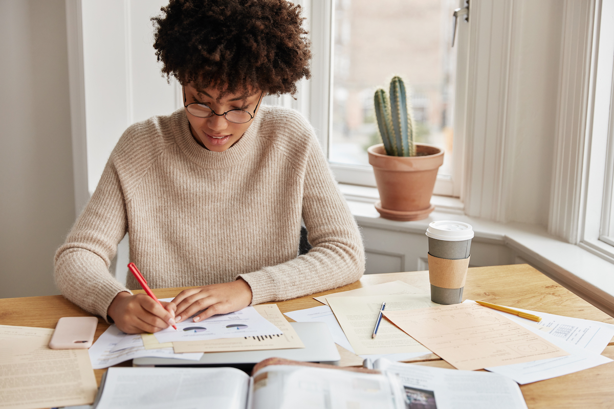 Busy accountant with curly dark hair, dressed casually, works on financial report at home, writes graphics and schemes, sits at desktop with coffee, documents and cell phone