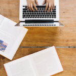A person works on a laptop on a wooden desk with two open books on it.