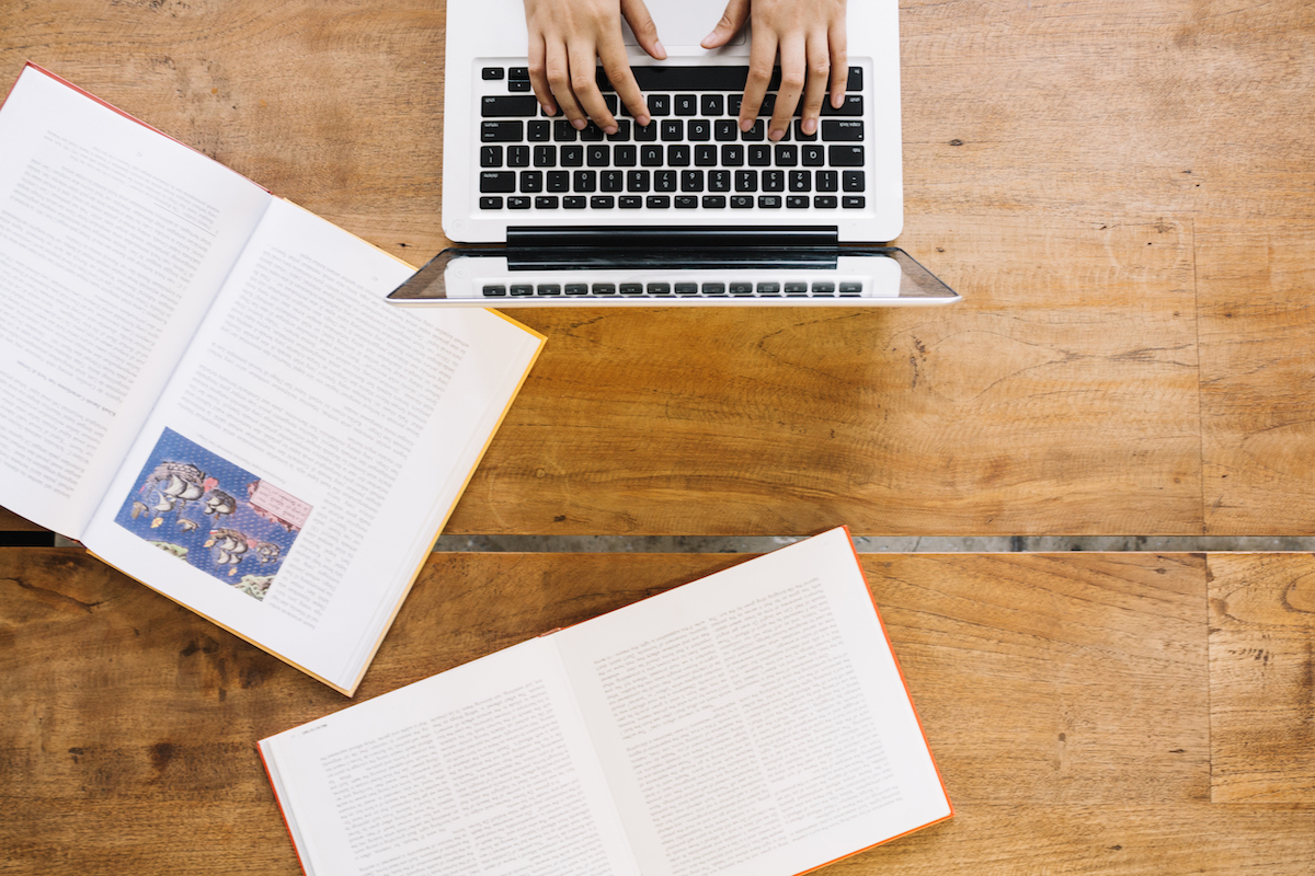 A person works on a laptop on a wooden desk with two open books on it.