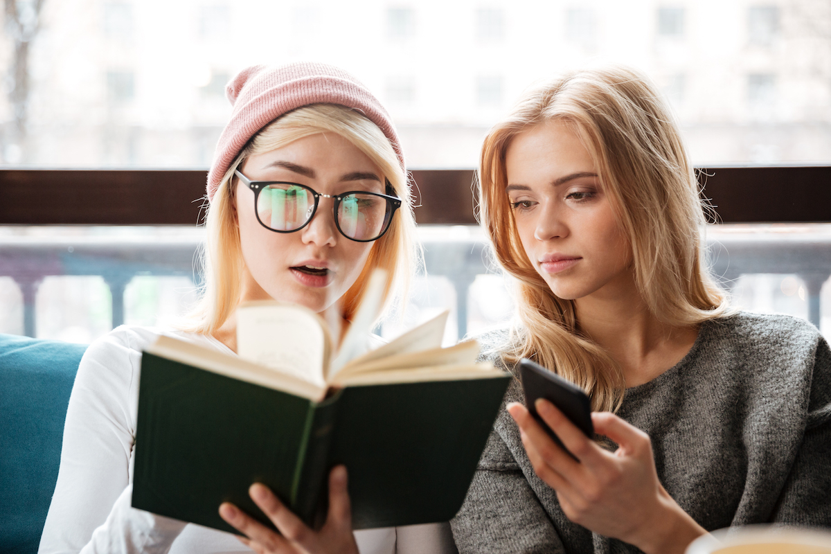 Image of two friends sitting in cafe and reading a book while holding phone