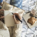 A person writes in a notebook on a blanket set out with snacks and a drink