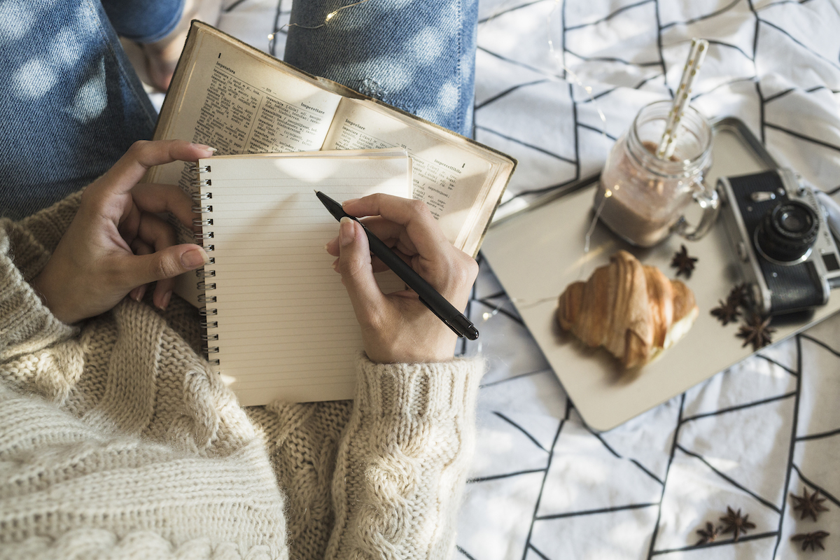 A person writes in a notebook on a blanket set out with snacks and a drink